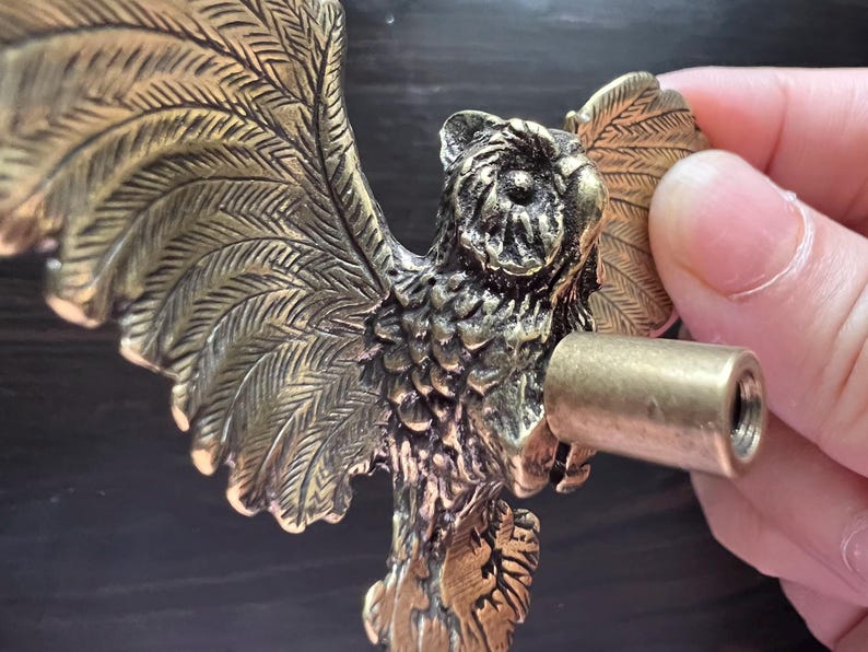 K&ouml;nnte beinhalten: Ein detaillierter, antik-messingfarbener Schrankgriff in Eulenform. Die Eule hat ausgebreitete Fl&uuml;gel mit detaillierten Federn. Ein zylindrischer, messingfarbener Schraubpfosten wird in der N&auml;he des K&ouml;rpers der Eule gehalten, bereit zur Installation. Die Hardware ist f&uuml;r M&ouml;bel konzipiert.