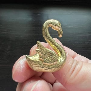 May include: A gold-colored swan figurine with detailed feather and wing textures. The swan has a curved neck and is held in a person's hand. The figurine is made of a shiny, metallic material.