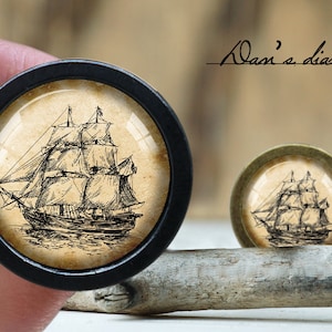 Può includere: Due cabochon rotondi con un tema nautico vintage. Uno è nero, l'altro è bronzo. Ognuno presenta un dettagliato schizzo in bianco e nero di una nave a vela su uno sfondo simile a pergamena. Il testo "Alan's diary" è visibile.