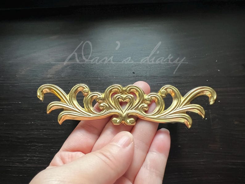 May include: Gold-toned metal decorative trim with a floral design featuring two hearts in the center. The trim has a delicate, ornate style.