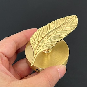 May include: A gold-colored feather-shaped object attached to a round base. The feather has detailed veining, and the base is a solid, circular form. The object is held against a dark background.