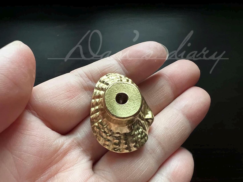 May include: A gold-colored metal cabinet knob with a decorative, textured design. The knob has a round, flat top with a small hole in the center.