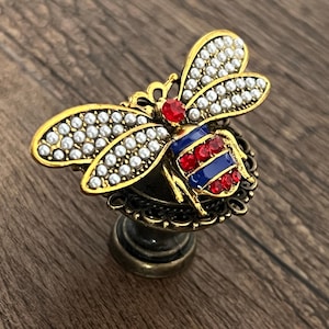 Puede incluir: Pomo de armario decorativo con forma de abeja, con detalles dorados, rojos, blancos y azules. Las alas de la abeja están adornadas con pequeñas perlas blancas. Montado sobre una base decorativa, ideal para muebles.
