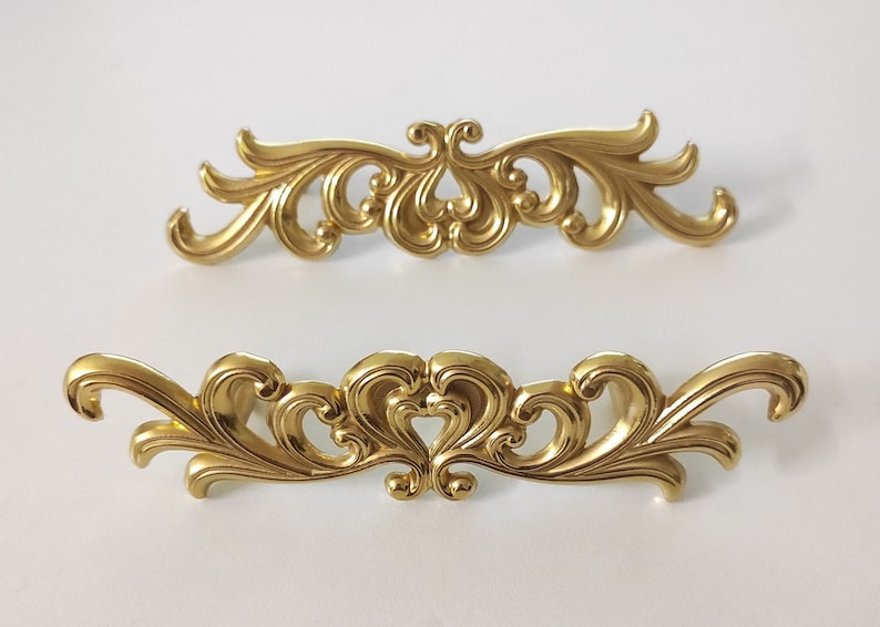 May include: Two ornate gold-toned metal drawer pulls with a decorative scroll design.