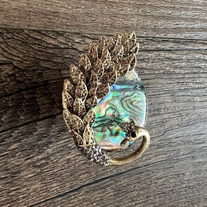 May include: A decorative brooch featuring a teardrop-shaped abalone shell with iridescent colors of blue, green, and gold. The shell is framed by a gold-colored metal design of leaves and a swan-like figure. The brooch is set against a wooden background.