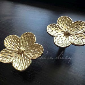 以下が含まれることがあります： テクスチャ加工された表面を持つ、金色の金属製のフラワー型の引き出しノブが2つあります。