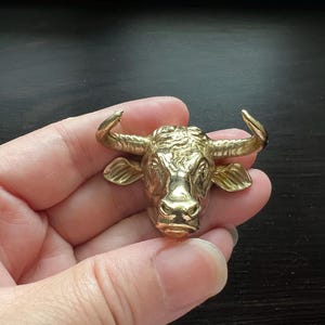 May include: A gold-coloured metal bull's head sculpture with detailed features. The sculpture is held in a person's hand.