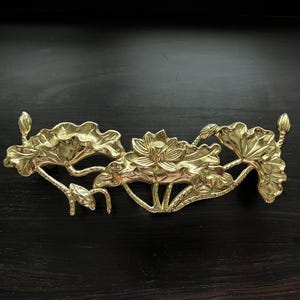 May include: A gold-coloured metal lotus flower design. The design is made up of three lotus flowers with leaves and stems. The design is intricate and detailed.