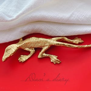 Peut inclure: Une figurine de lézard en métal doré sur une surface rouge. Le lézard est en position de course, la queue enroulée.
