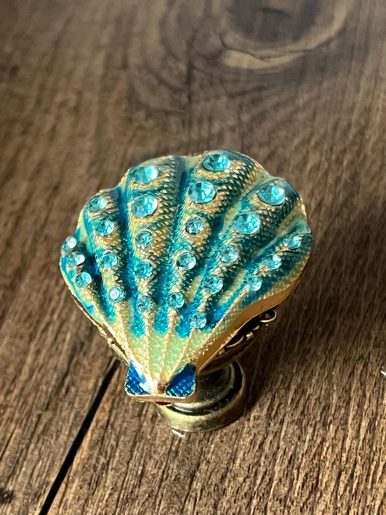 May include: A decorative seashell-shaped knob with a teal and gold finish. The knob is adorned with teal-coloured rhinestones and has a brass-coloured base. The seashell design is detailed and eye-catching, perfect for adding a touch of coastal charm.
