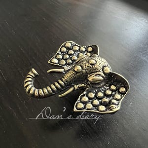 Peut inclure: Broche éléphant de style antique en bronze. La tête et les oreilles de l'éléphant sont ornées de petits détails circulaires en relief. La broche est posée sur un fond sombre et texturé, avec le texte "Dan's diary" en cursive.