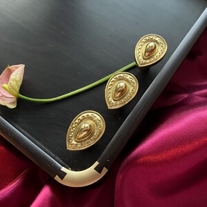 May include: Three gold-toned decorative drawer pulls in a teardrop shape with a textured design. The pulls are on a black surface.