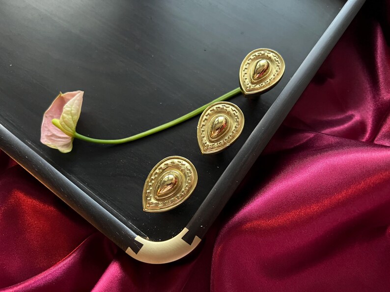 May include: Three gold-toned decorative drawer pulls in a teardrop shape with a textured design. The pulls are on a black surface.