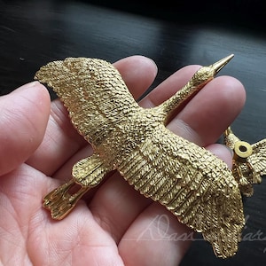 Op de afbeelding: Een gouden metalen kraanvogelfiguur met gedetailleerde vleugels en een lange nek. De kraanvogel is in vlucht en heeft een realistisch ontwerp.