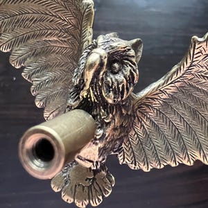 K&ouml;nnte beinhalten: Eine detaillierte Eulen-Skulptur aus Messing mit ausgebreiteten Fl&uuml;geln und einem zylindrischen Lauf, der aus der Brust ragt. Die Eule ist aufwendig mit strukturierten Federn und einem fokussierten Blick versehen.