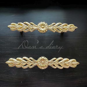May include: Two gold-toned decorative drawer pulls with a floral design. Each pull features a central flower with intricate details and flowing leaves.