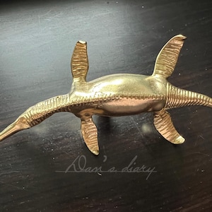 May include: A gold-coloured brass sculpture of a prehistoric marine reptile, resembling a plesiosaur, with a long neck and flippers. The sculpture is detailed with textured scales and fins.