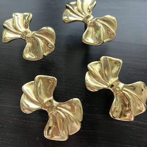 May include: Four gold-toned metal bow-shaped drawer pulls. The pulls are all the same design, but are oriented in different directions.