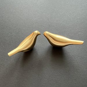 Pu&ograve; includere: Due statuine di uccelli in metallo color oro. Gli uccelli si guardano, con i loro becchi che si toccano. Gli uccelli sono su uno sfondo grigio scuro.