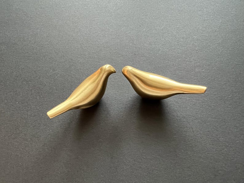 Pu&ograve; includere: Due statuine di uccelli in metallo color oro. Gli uccelli si guardano, con i loro becchi che si toccano. Gli uccelli sono su uno sfondo grigio scuro.