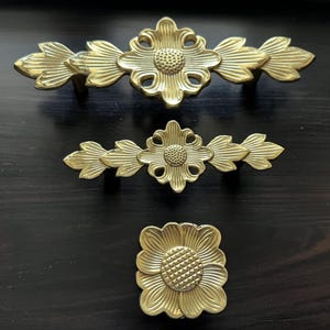 Könnte beinhalten: Drei goldfarbene Metall-Schubladengriffe mit floralen Designs. Der obere Griff hat eine größere Blume in der Mitte mit zwei kleineren Blumen auf jeder Seite. Der mittlere Griff hat eine kleinere Blume in der Mitte mit zwei kleineren Blumen auf jeder Seite. Der untere Griff ist eine einzelne Blume.