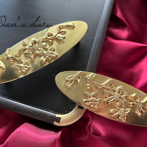 Pode incluir: Dois grampos de cabelo dourados com um design floral. Os grampos estão sobre uma superfície preta com uma superfície vermelha no fundo.