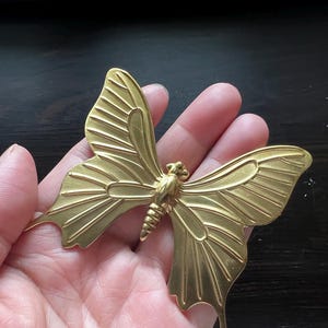 May include: A gold-colored metal butterfly ornament with detailed wings. The butterfly is held in a hand, but the hand is not visible in the image.