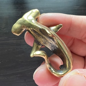 Puede incluir: Un llavero de latón con forma de tiburón martillo. El tiburón tiene aletas detalladas y una cola curvada, con un lazo para llaves. El metal tiene un acabado dorado pulido.