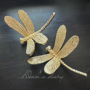 May include: Two gold-coloured dragonfly decorative objects. The dragonflies have detailed wings and bodies, with a textured surface. The objects are set against a dark background. The dragonfly's wings are spread out.