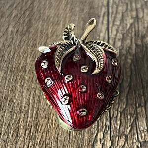 Op de afbeelding: Een decoratieve aardbeivormige sieradendoos. De doos is dieprood van kleur met gouden accenten, waaronder de steel en kleine zaaddetails. De doos is gemaakt van metaal en heeft een scharnierende opening.