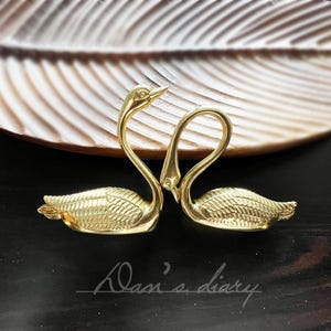 May include: Two gold swan-shaped decorative pieces, facing each other, with their necks intertwined. The swans are on a brown, textured surface.