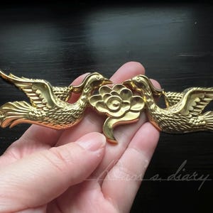 Puede incluir: Pieza decorativa de metal dorado con dos pájaros estilizados que se miran, con un diseño floral central. Los pájaros tienen alas y picos detallados, y la flor tiene un centro circular y cinco pétalos.
