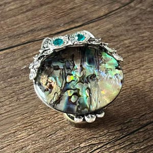 Può includere: Un portagioie decorativo con una cornice in metallo argentato e un top rotondo in conchiglia di abalone iridescente. La parte superiore è adornata con filigrana argentata e gemme color turchese. La scatola poggia su una superficie di legno.