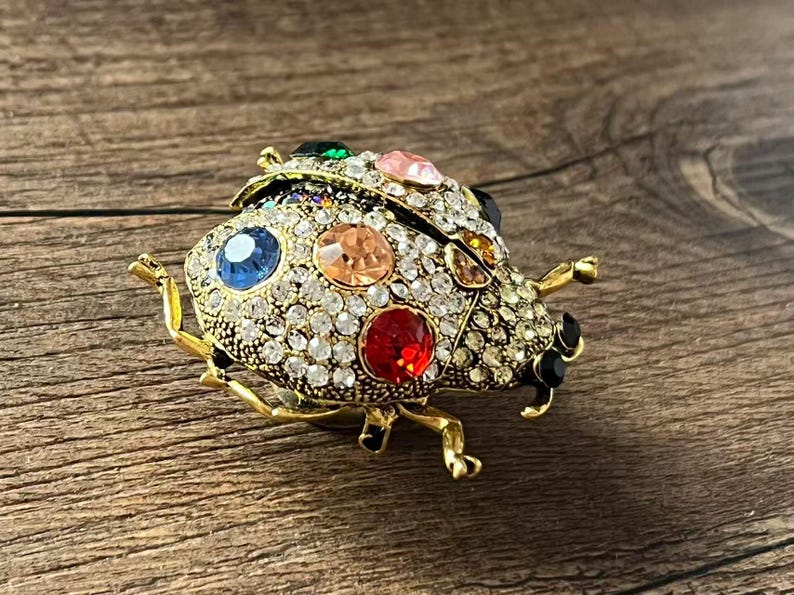 May include: A gold-coloured ladybird brooch adorned with clear and colourful gemstones. The brooch has a detailed design with a mix of round and oval-shaped gems in various colours, including red, blue, and pink. The ladybird's body is covered in small, clear crystals.