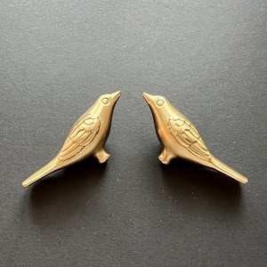 Puede incluir: Dos figuritas de pájaros de metal dorado que se miran. Los pájaros tienen alas y picos detallados.