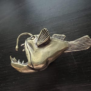 May include: A bronze sculpture of a anglerfish with a large mouth and a lure on its head. The sculpture is detailed and realistic.