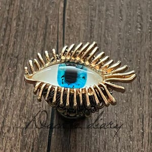 May include: A gold-colored eye-shaped pin with a blue iris and white sclera. The eye is surrounded by gold-colored eyelashes. The pin is set against a wooden background.