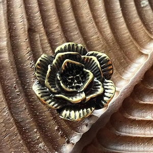 May include: A detailed, antique-style brass rose-shaped knob. The knob is a dark gold color with intricate petal detailing. It is set against a textured, brown wooden surface, creating a contrast in color and texture. This decorative hardware is suitable for furniture or cabinetry.