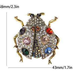 May include: A gold-coloured ladybug brooch, 5.8 cm long and 4.3 cm wide, adorned with clear and colourful gemstones. The brooch has a detailed design with multiple coloured gems and a gold-coloured metal frame.
