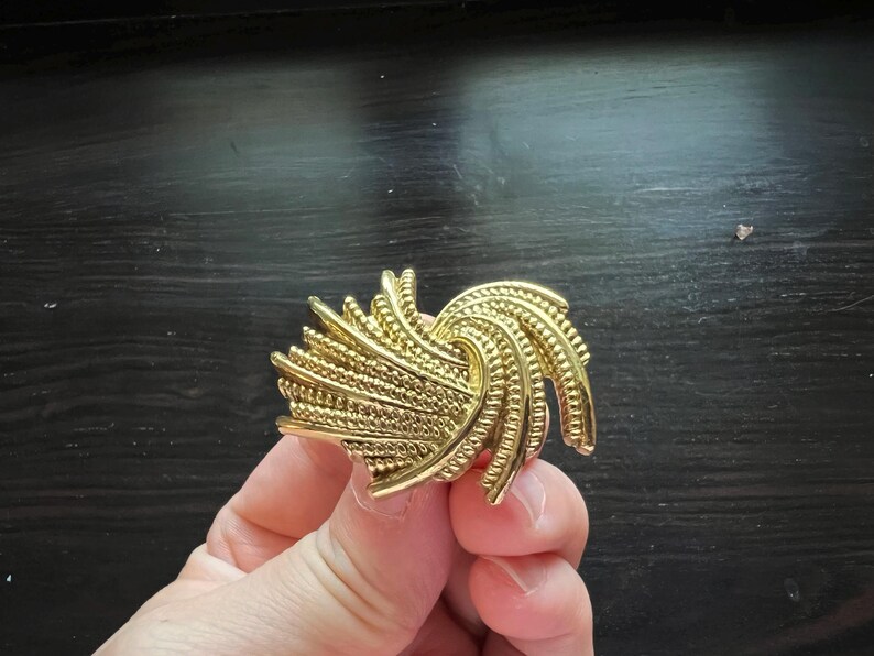 K&ouml;nnte beinhalten: Eine goldfarbene Vintage-Brosche mit einem wirbelnden, strukturierten Design. Die Brosche hat eine Reihe von gebogenen, geriffelten Elementen und ein strukturiertes, perlenbesetztes Muster. Das Schmuckst&uuml;ck wird in einer Hand vor dunklem Hintergrund gehalten.