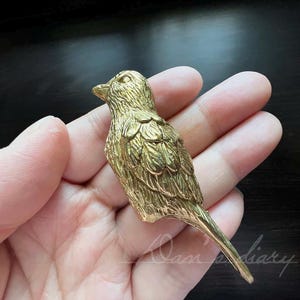 Op de afbeelding: Een gouden metalen vogelbeeldje met gedetailleerde veren. De vogel zit op een hand.