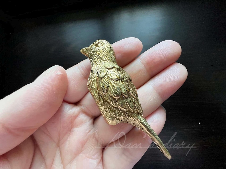 Puede incluir: Una figurita de p&aacute;jaro de metal dorado con plumas detalladas. El p&aacute;jaro est&aacute; posado en una mano.
