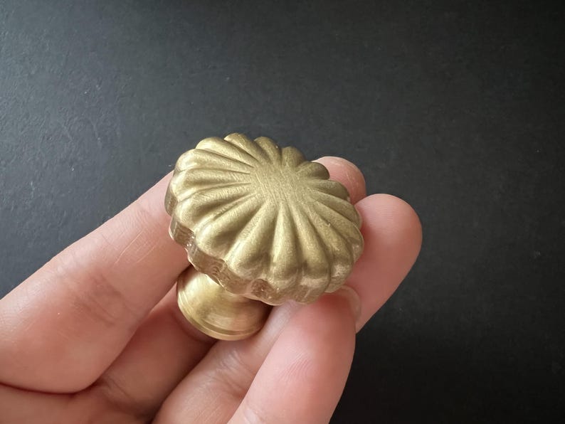 Puede incluir: Un pomo de armario de metal dorado con un dise&ntilde;o acanalado y festoneado. El pomo tiene una base redonda y una parte superior decorativa elevada.