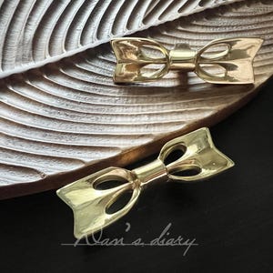 May include: Two gold-coloured bow-shaped decorative objects. The bows are placed on a wooden, leaf-shaped tray. The bows have a polished finish and are positioned on a dark surface. The text "Nasi's diary" is written in cursive.