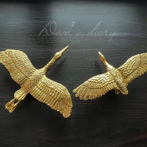 May include: Two gold-colored metal crane sculptures with outstretched wings. The cranes are facing opposite directions.