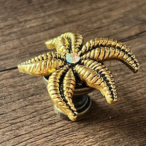 Puede incluir: Un objeto decorativo dorado en forma de estrella de mar con una gema iridiscente central. La estrella de mar tiene crestas detalladas y está montada sobre una pequeña base decorativa. Probablemente un tirador de armario.