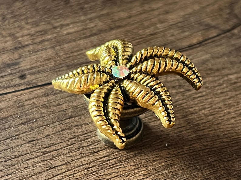 Op de afbeelding: Een goudkleurig, stervormig decoratief object met een centrale iriserende edelsteen. De zeester heeft gedetailleerde ribbels en is gemonteerd op een kleine, decoratieve basis. Waarschijnlijk een kastknop.