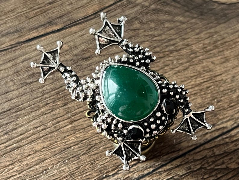 以下が含まれることがあります： 中央に大きな楕円形の緑色の宝石が付いた、詳細なカエル型のブローチ。ブローチは、テクスチャ加工された脚やスタッドボディなど、複雑なディテールを備えたシルバー調の仕上げです。カエルは木製の表面に配置されています。