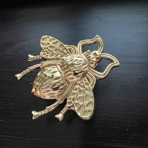 Può includere: Un fermaglio a forma di ape color oro. Il fermaglio presenta ali, segmenti del corpo e zampe dettagliate. L'ape è posta su uno sfondo scuro, evidenziando la sua lucentezza metallica. Questa spilla decorativa è adatta per giacche, borse o cappelli.
