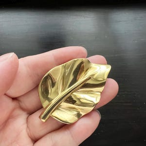 May include: A gold-toned leaf-shaped brooch. The brooch is made of metal and has a detailed design.